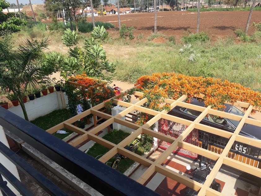 A pergola grid built over the courtyard and car porch of an existing house, with a flowering creeper trained across it